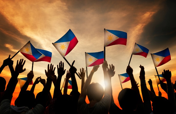 A crowd's silhouetted hands hold up Phillipines flags against a golden sunset
