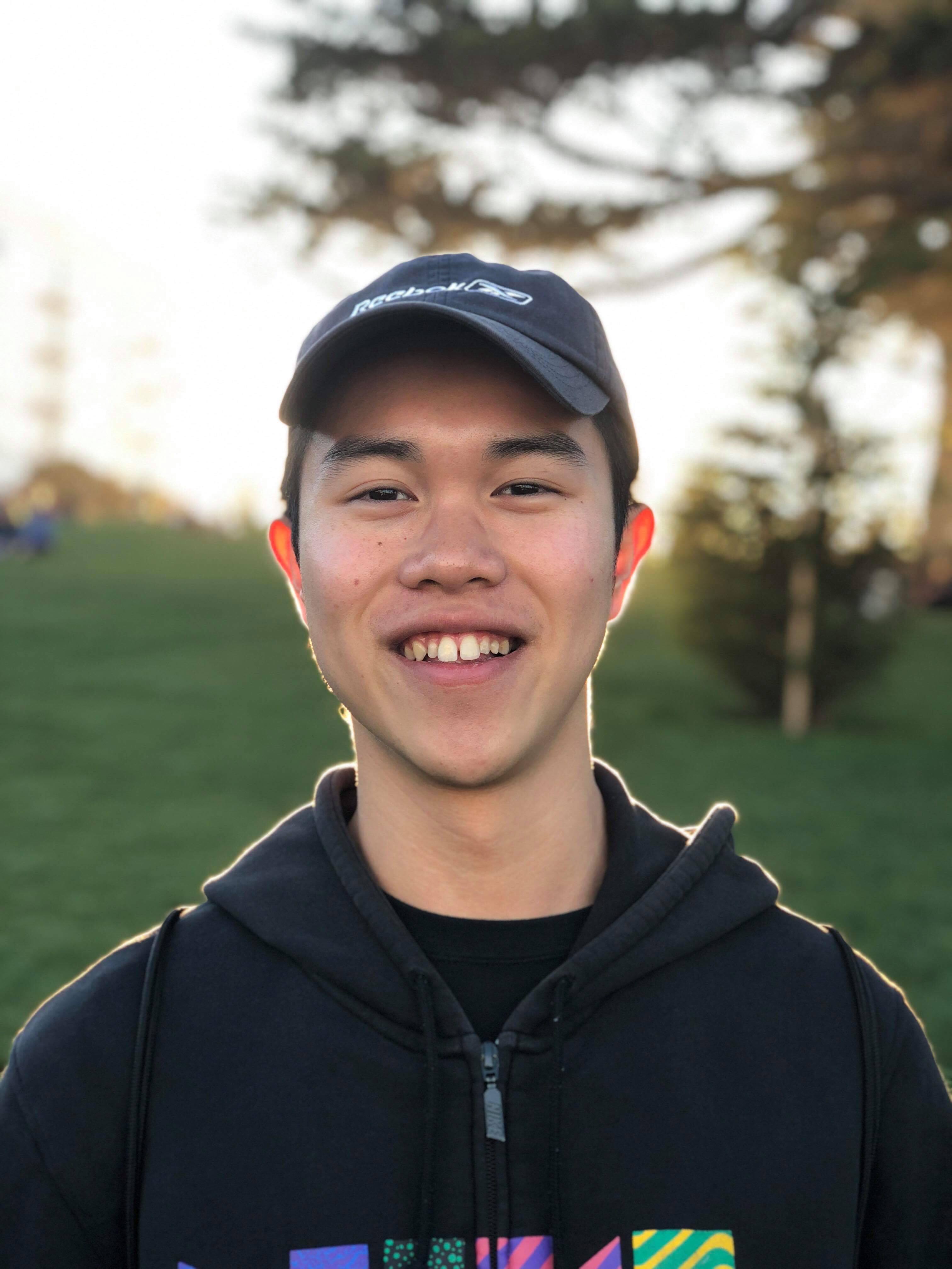 Smiling young student in a black cap and hoodie standing in a grassy park at sunset.