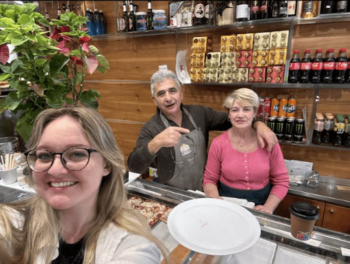blonde girl takes a selfie with a smiling couple standing behind a glass pastry case in an Italian cafe