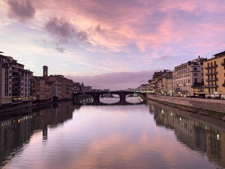 a view of the Ponte Vecchio bridge during a pink and purple sunset reflecting off the sky and water