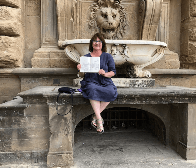 smiling woman holds up her sketchbook as she sits on an Italian stone fountain