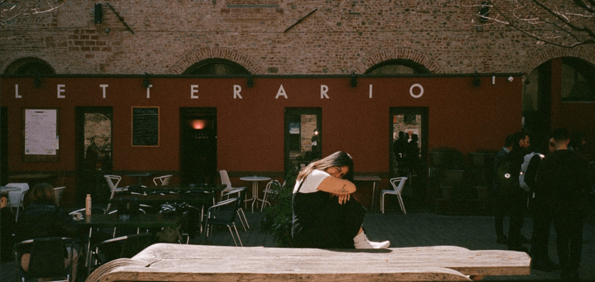 a girl in black sits in the sunlight on a wooden art piece in front of a shaded Italian courtyard cafe